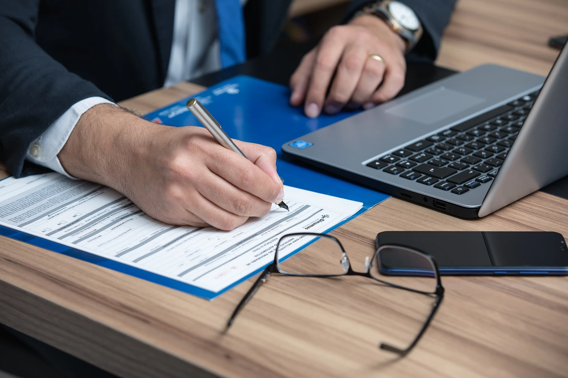 A person writing a report by their open lap top with a phone and glasses on the table.