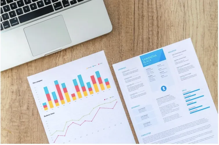 Two sheets of paper with data on the sheets and a lap top all on a table.